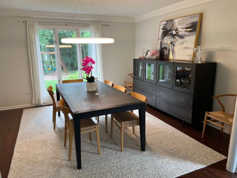 A dining room with a dark wood table, six wooden chairs, a buffet cabinet, modern artwork, and a large window overlooking a backyard. A potted orchid is centered on the table.
