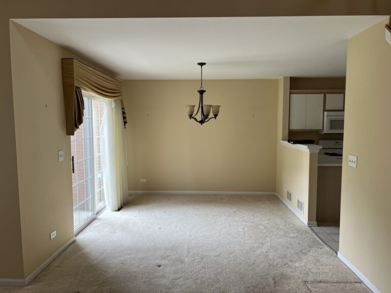 Empty dining area with beige walls and carpet, a ceiling light fixture, sliding glass door with curtains, and partial view of a kitchen with white cabinets and appliances.
