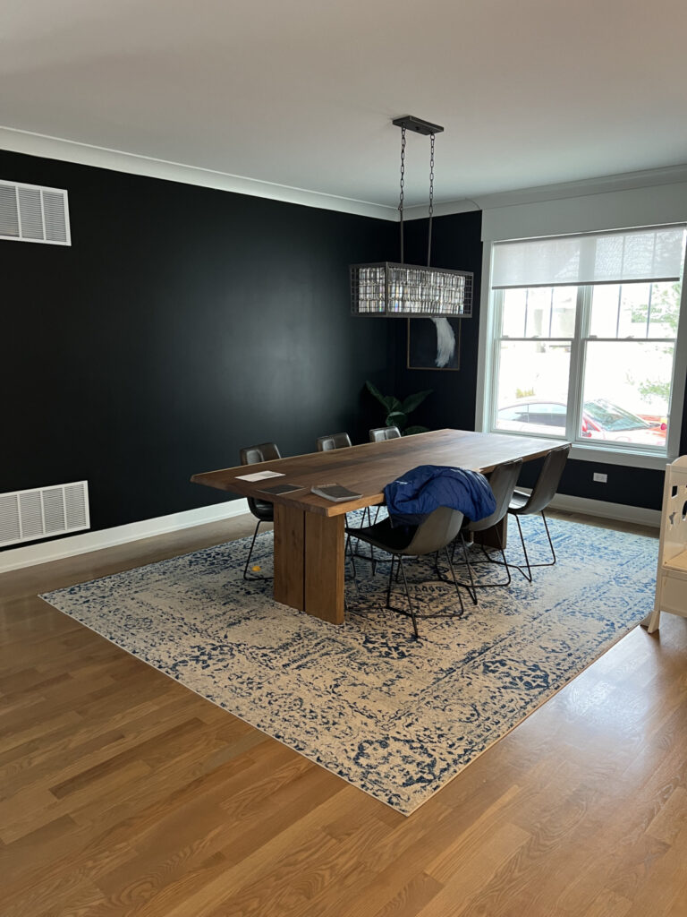A dining room with a large wooden table, six chairs, a blue jacket draped over a chair, a patterned rug, black accent wall, and a window with natural light.