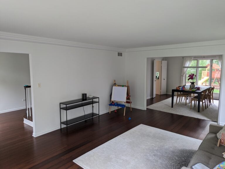 A spacious living and dining area with hardwood floors, white walls, a small black console table, easel, dining table, and large windows showing a backyard playset.
