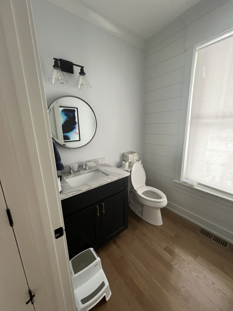 A modern bathroom with a black vanity, round mirror, sink, toilet, toilet paper, step stool, wall art, and a large window with a shade.