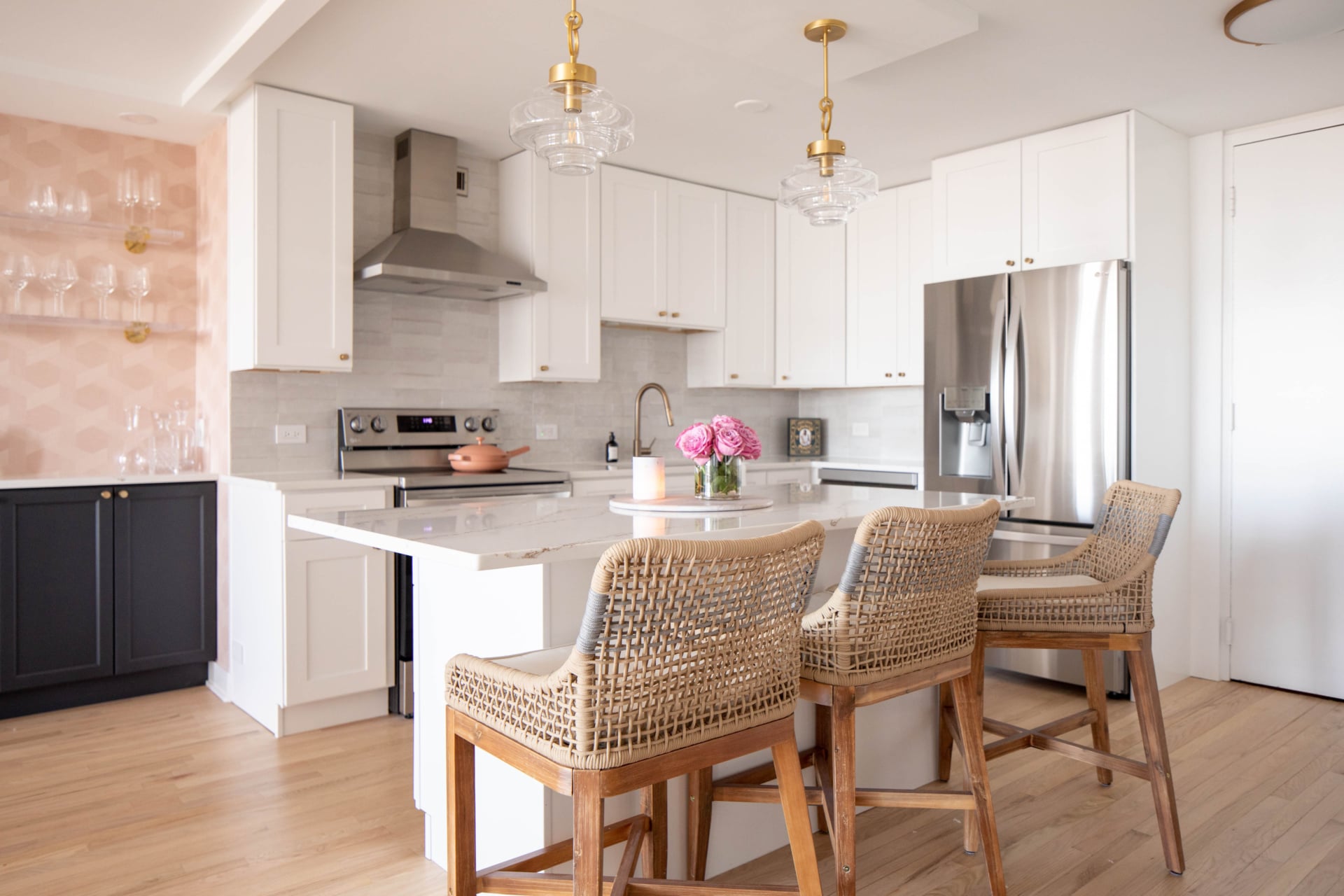 Modern kitchen with white cabinets, stainless steel appliances, a central island with two wicker barstools, and a vase of pink flowers on the countertop.