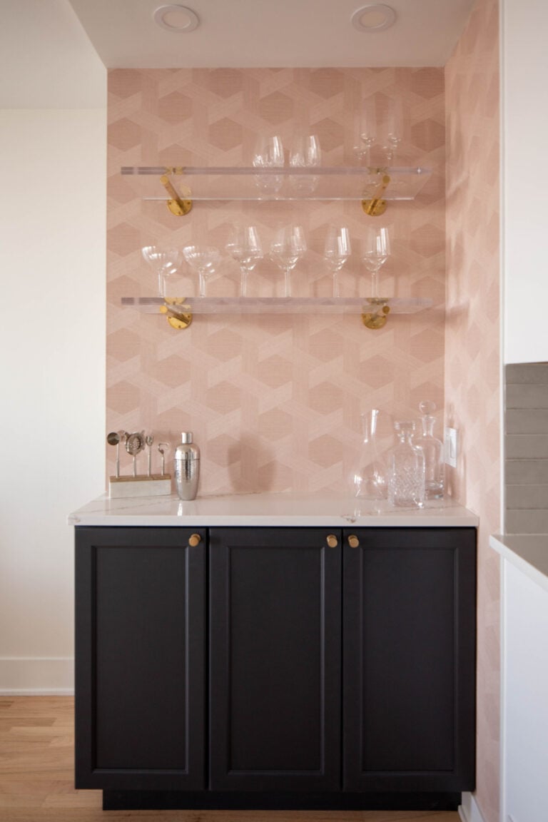 A home bar area with black cabinets, a white countertop, glassware, and decanters on top; two glass shelves with glasses are mounted on a pink geometric wallpapered wall.