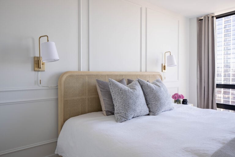 A neatly made bed with a rattan headboard, gray patterned pillows, white bedding, wall-mounted lamps, and a window with city view and light curtains.