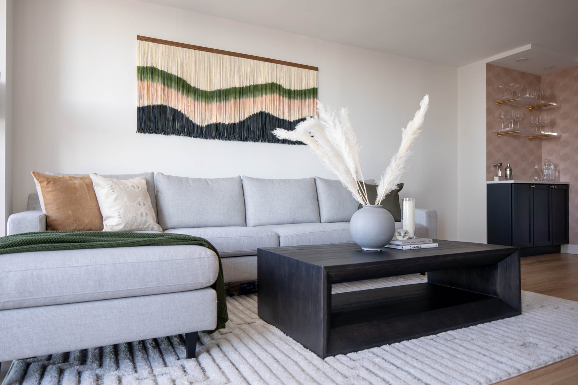Modern living room with a light gray sectional sofa, black coffee table, decorative vase with pampas grass, wall art, and a small bar area in the background.
