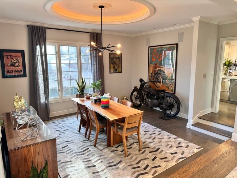 A dining room with a wooden table and six chairs, large window with gray curtains, framed posters, a vintage motorcycle, modern light fixture, and an accent rug on a hardwood floor.