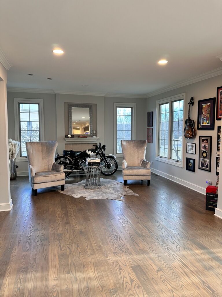 A modern living room with two high-back chairs, a small glass table, a motorcycle on display, framed art on the walls, and a guitar hanging next to large windows.