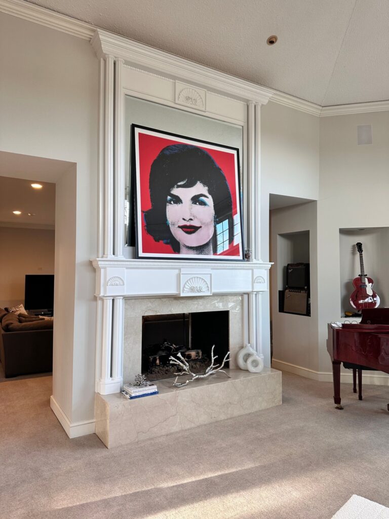A white fireplace with a large pop art portrait of a woman above it, a red guitar on a stand, and a red grand piano in the living room.