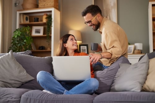 A woman sits on a couch with a laptop on her lap, smiling up at a man standing beside her holding a mug. They appear to be having a casual conversation in a living room.