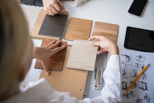 People examine and compare several wood laminate samples on a table with a sketch of furniture designs, a pencil, a tablet, and a clipboard.