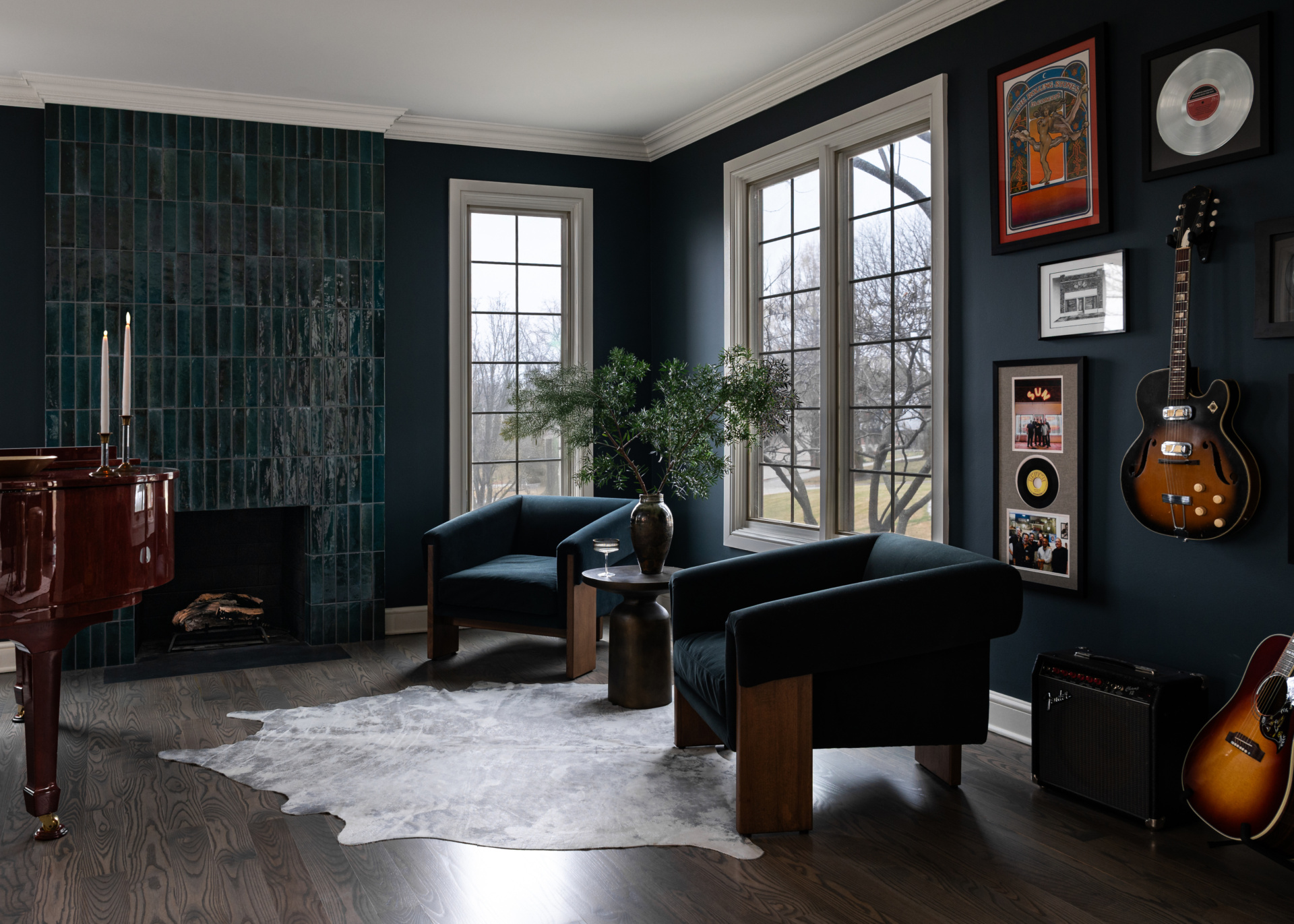 Dark, modern living room with two velvet armchairs, a tiled fireplace, a grand piano, wall art, a guitar, and large windows overlooking trees. A cowhide rug covers the wood floor.