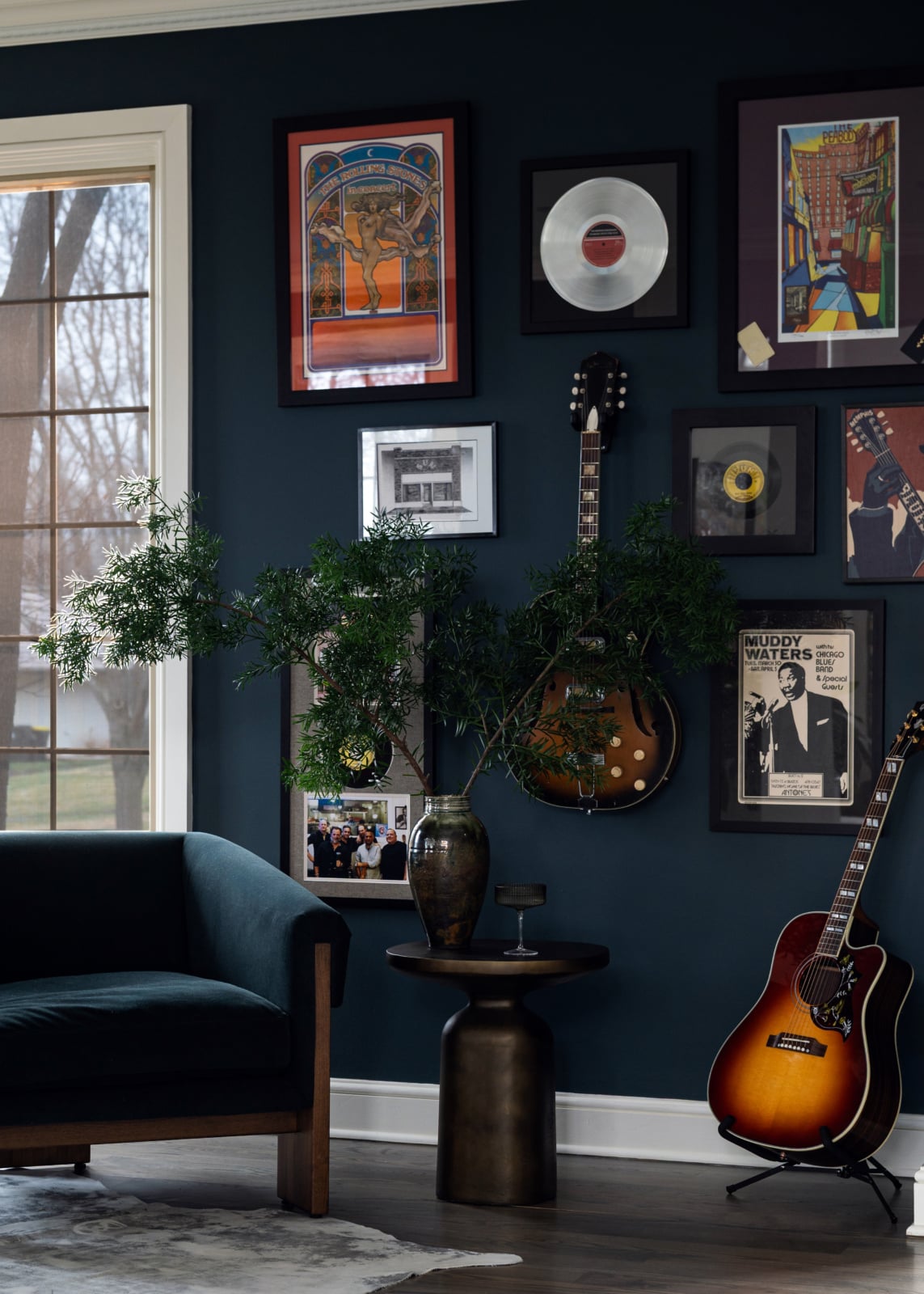 A dark blue wall decorated with framed art, records, two hanging guitars, a sunburst guitar on the floor, a potted plant on a round table, and a dark armchair by a window.