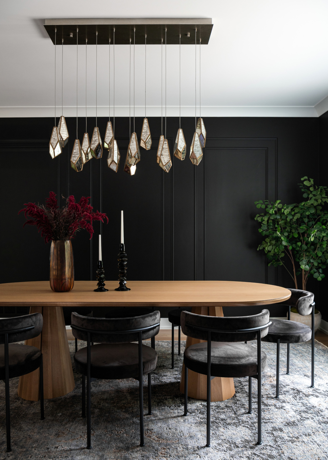 Modern dining room with a long wooden table, black chairs, a geometric chandelier, black paneled walls, a vase with dark red flowers, two candlesticks, and a potted plant.