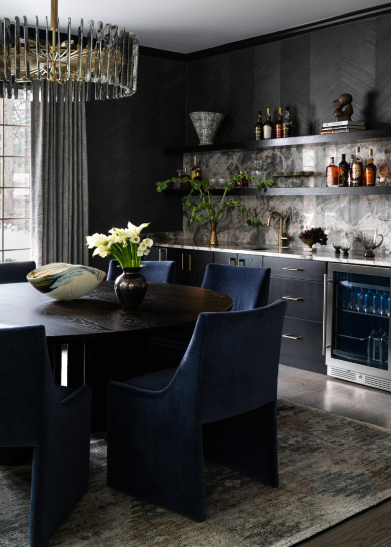 Modern dining room with dark walls, a round table, navy chairs, floral centerpiece, open shelves with bottles, marble backsplash, and a glass-front mini fridge.