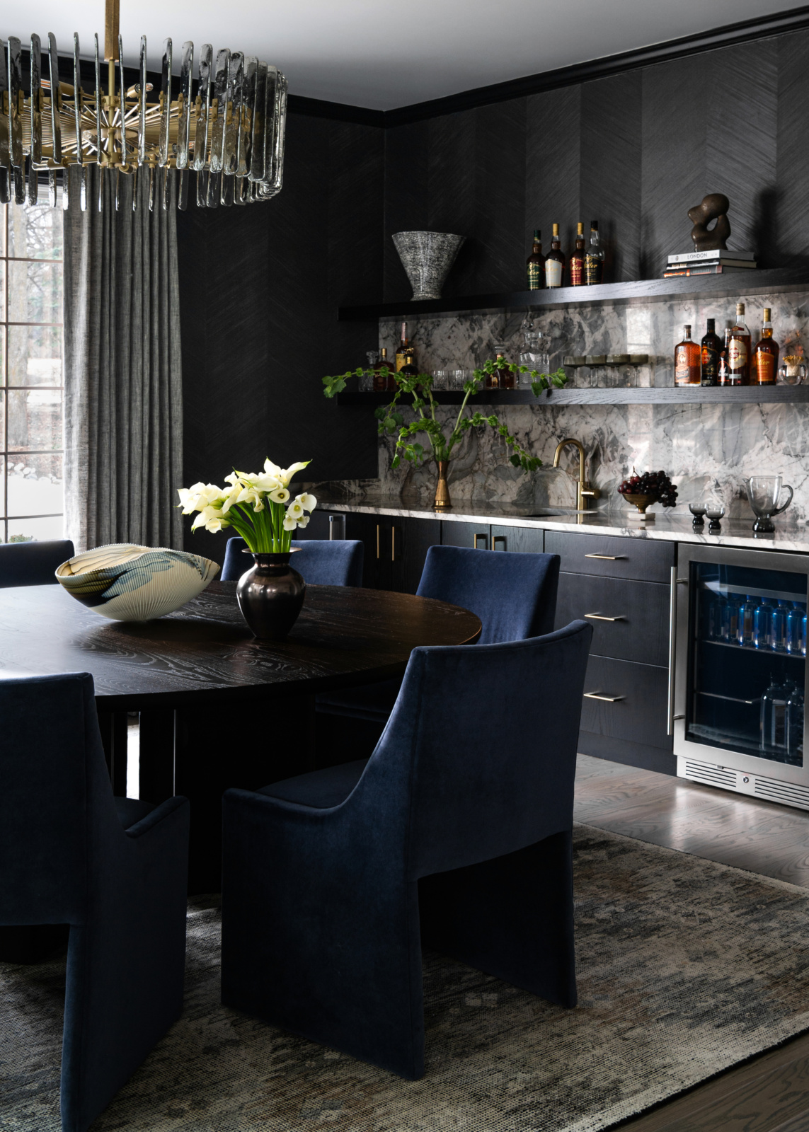 Modern dining room with dark walls, a round table, navy chairs, floral centerpiece, open shelves with bottles, marble backsplash, and a glass-front mini fridge.
