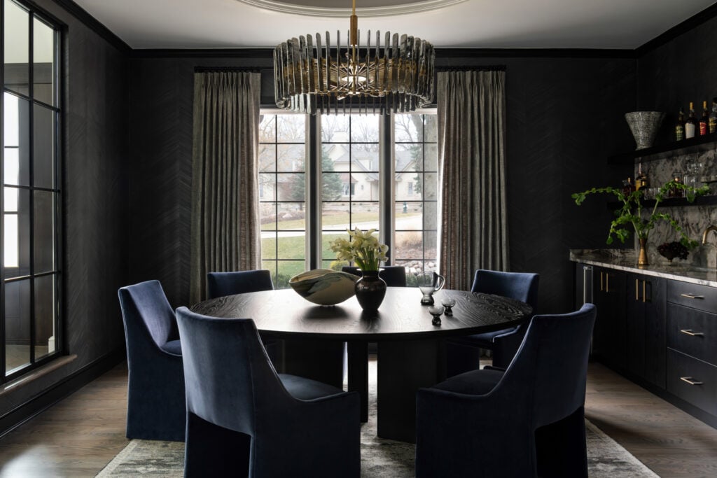 A modern dining room with dark walls, a round black table, six blue chairs, a chandelier, large windows with curtains, and a sideboard with plants and glassware.
