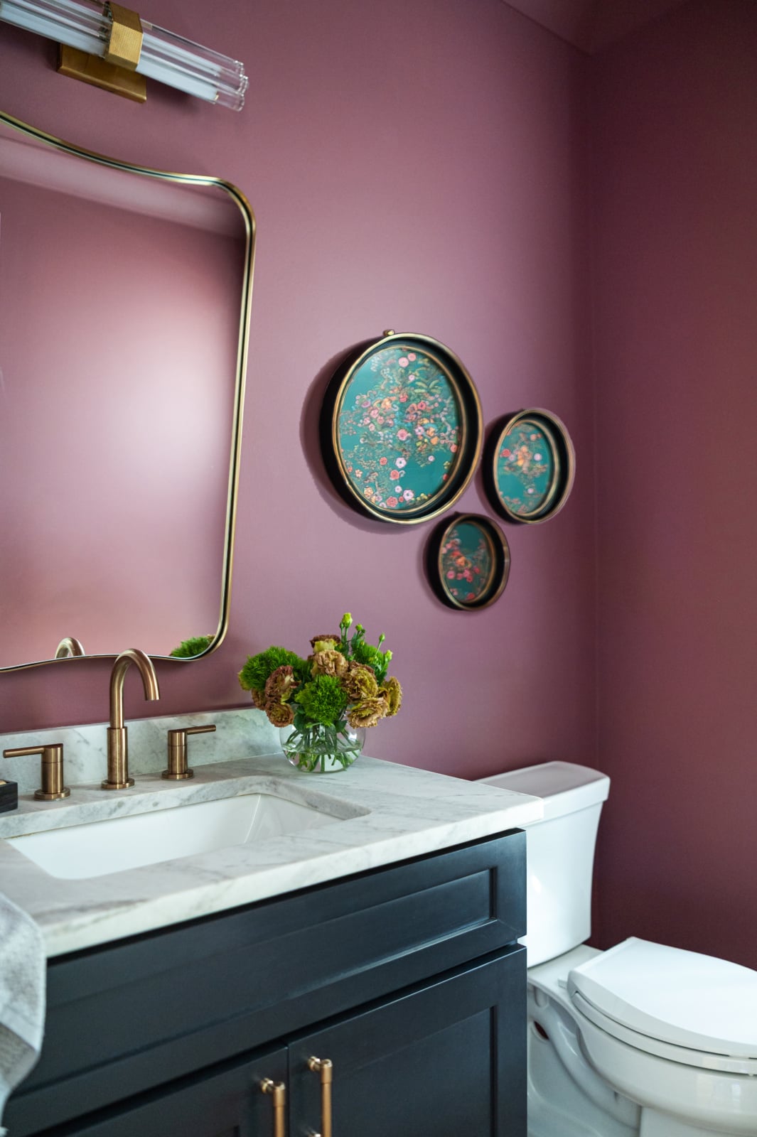 Bathroom with mauve walls, a marble countertop, gold fixtures, dark vanity, wall art in round frames, and a vase of flowers beside a white toilet.