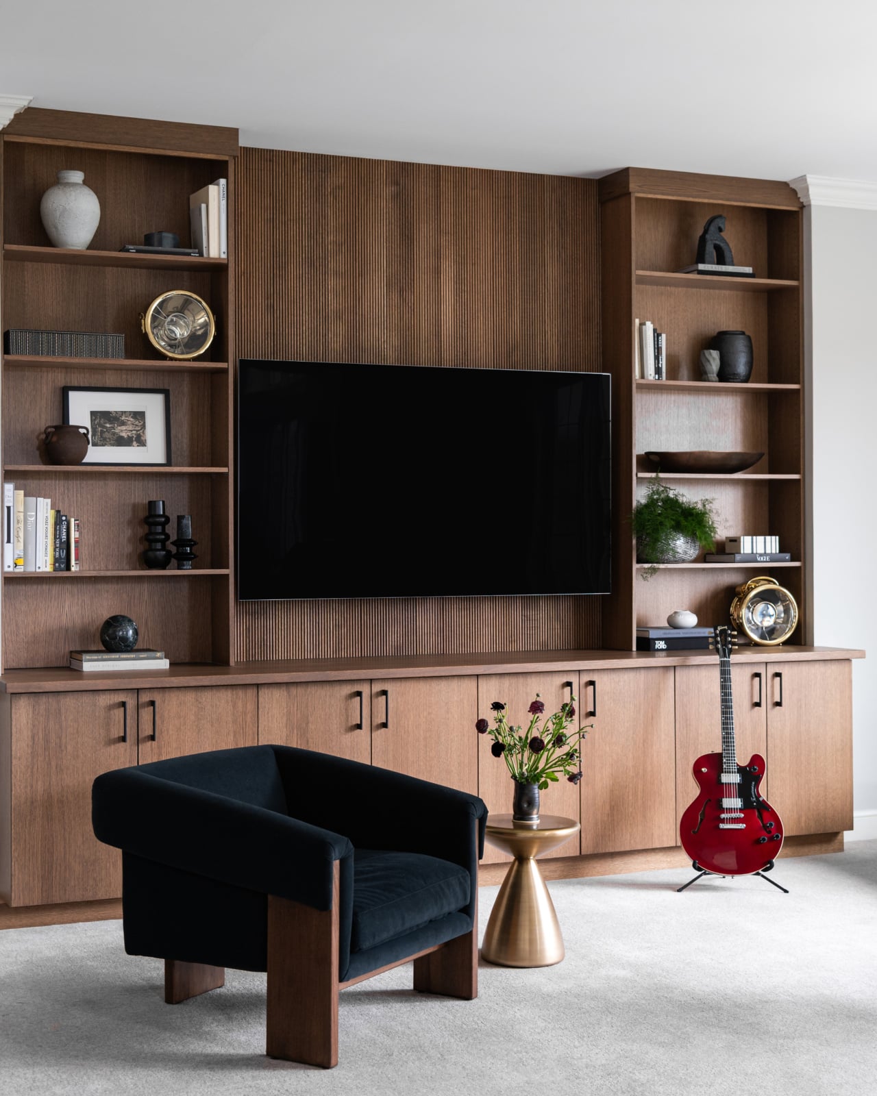 A modern living room with a wall-mounted TV, built-in wooden shelves, a black armchair, a small round table with flowers, and a red electric guitar on a stand.