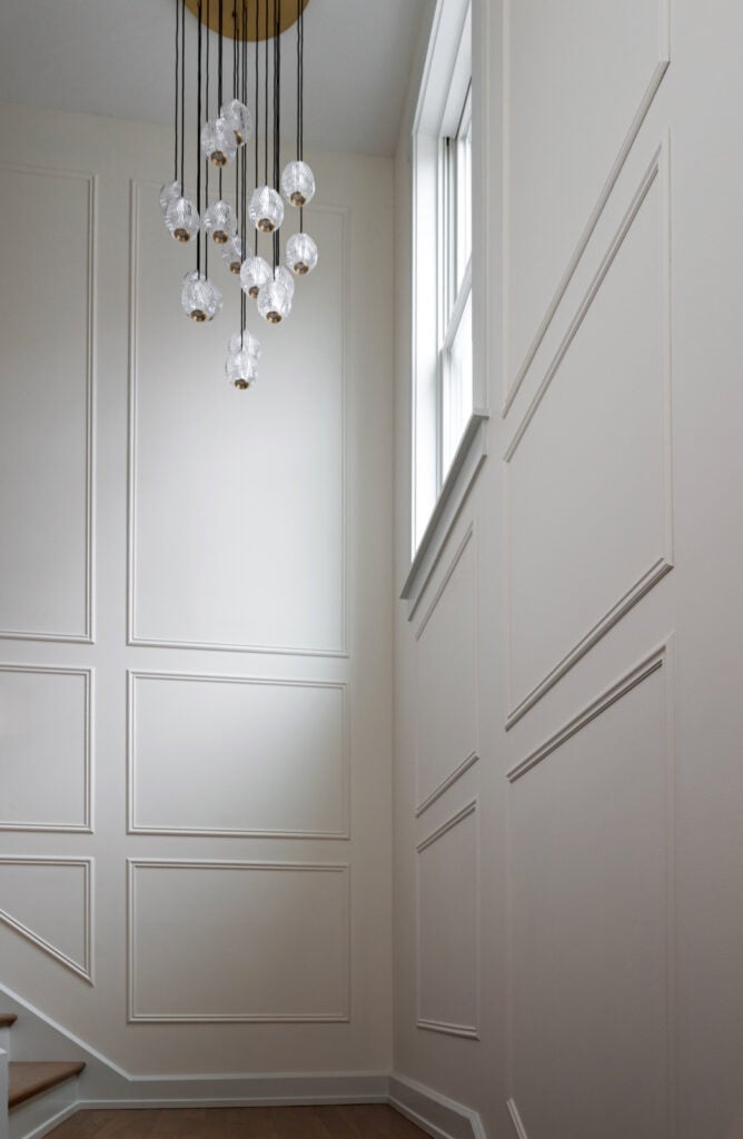 A modern staircase with white paneled walls, a large window, and a hanging chandelier with multiple glass globe lights.