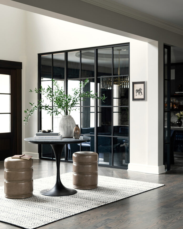 A modern entryway with a round table, two beige ottomans, a white vase with greenery, and a light rug on dark wood floors; glass doors and framed art are in the background.