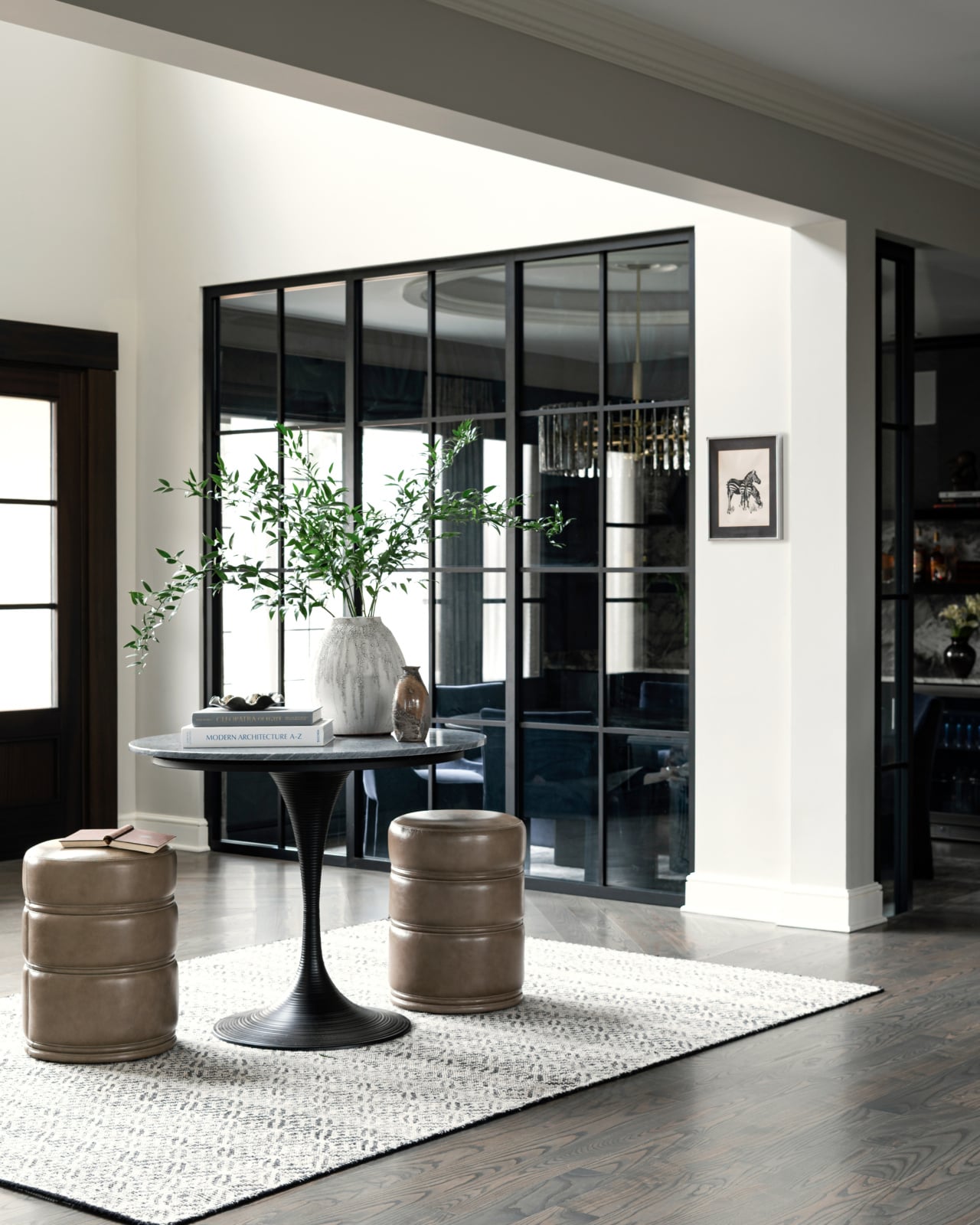 A modern entryway with a round table, two beige ottomans, a white vase with greenery, and a light rug on dark wood floors; glass doors and framed art are in the background.