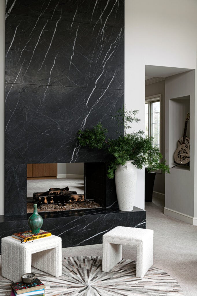 A modern living room with a black stone fireplace, a large white vase with greenery, two small textured stools, a round rug, and a guitar in the alcove.