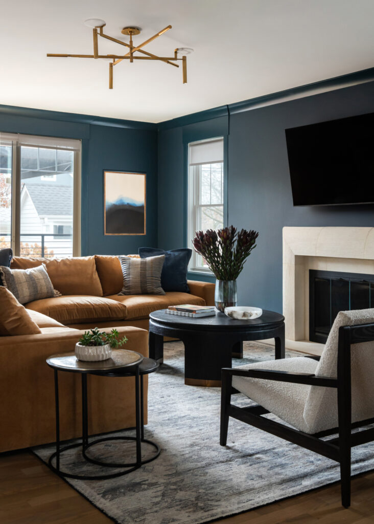 A modern living room with a tan sectional sofa, black coffee table, armchair, wall-mounted TV, fireplace, and large windows with blue walls.