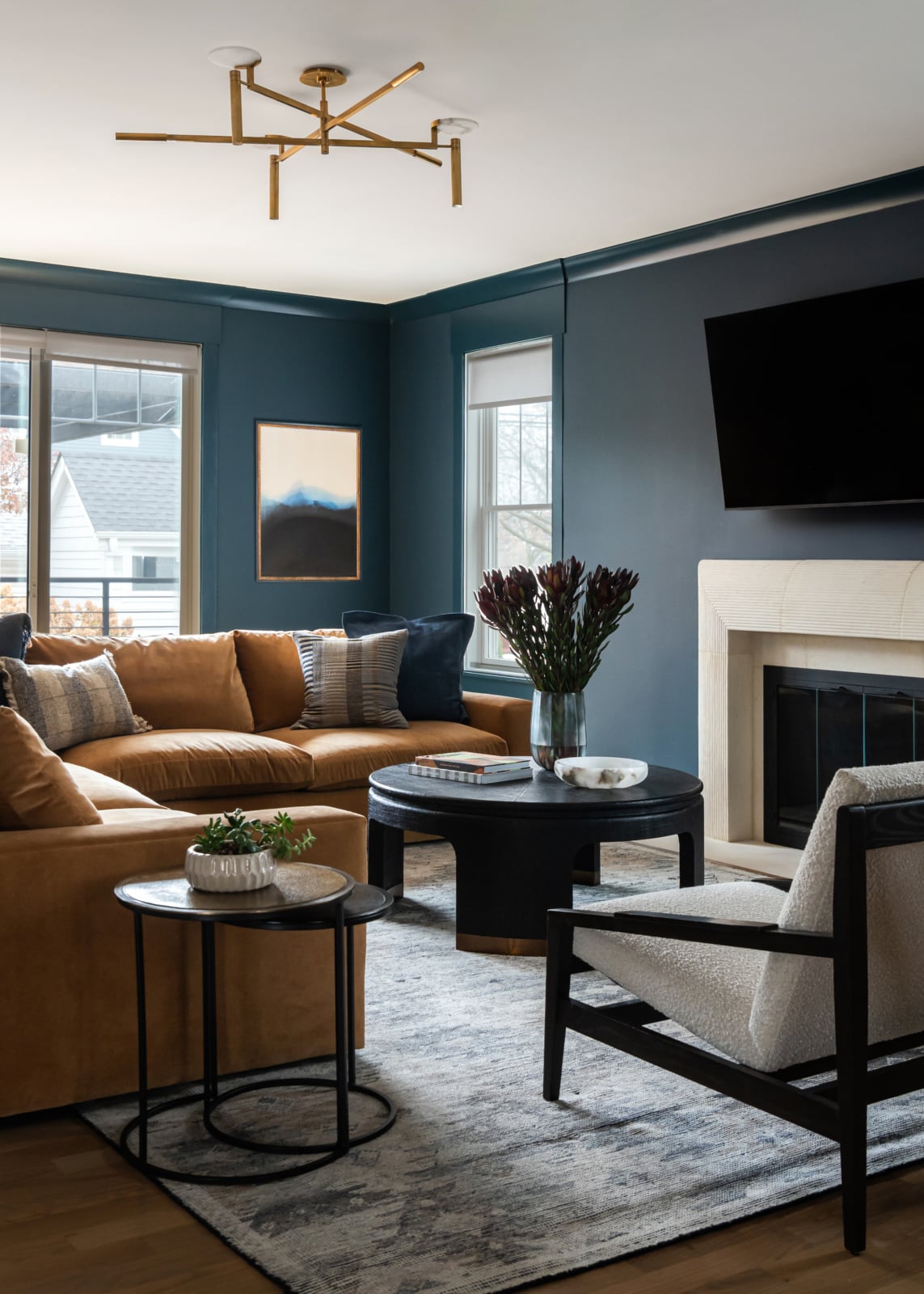 A modern living room with a tan sectional sofa, black coffee table, armchair, wall-mounted TV, fireplace, and large windows with blue walls.