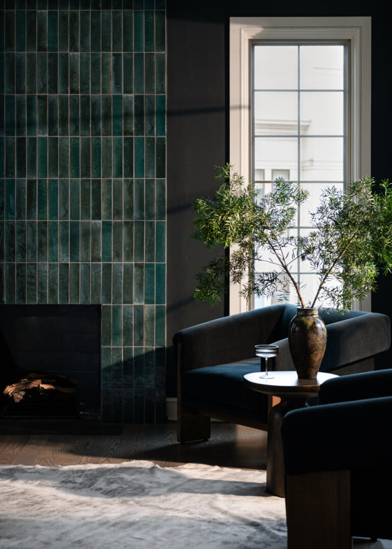 Modern living room with dark green tiled fireplace, two black armchairs, a round table with a large vase of greenery, and a window letting in natural light.