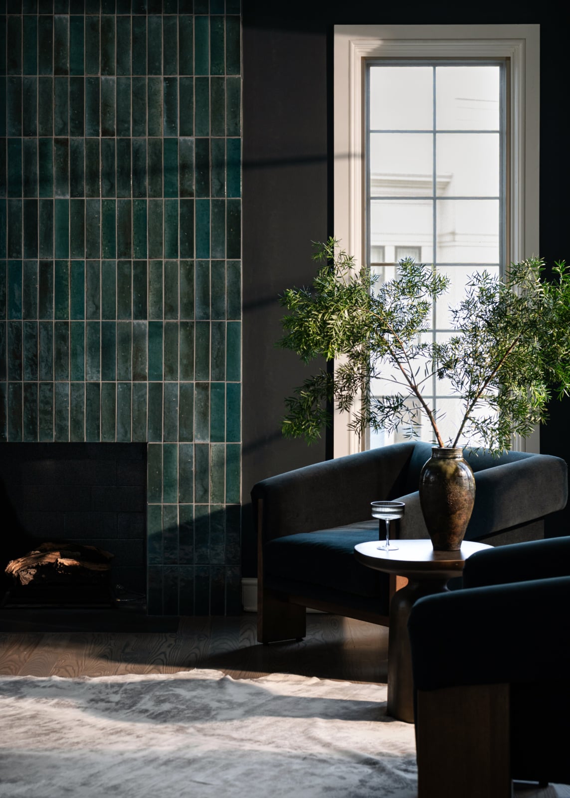 Modern living room with dark green tiled fireplace, two black armchairs, a round table with a large vase of greenery, and a window letting in natural light.