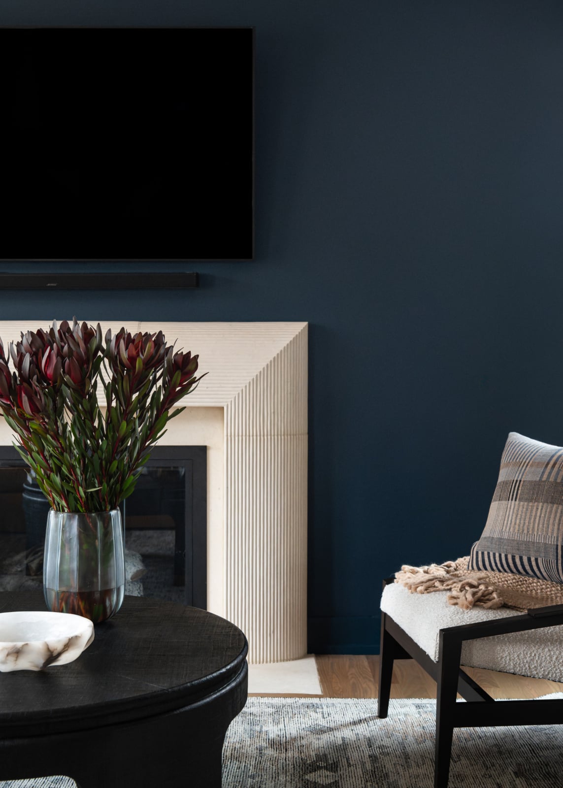 A modern living room with a dark blue wall, mounted TV, light-colored fireplace, armchair with cushions, round table, and a vase of dark red flowers.