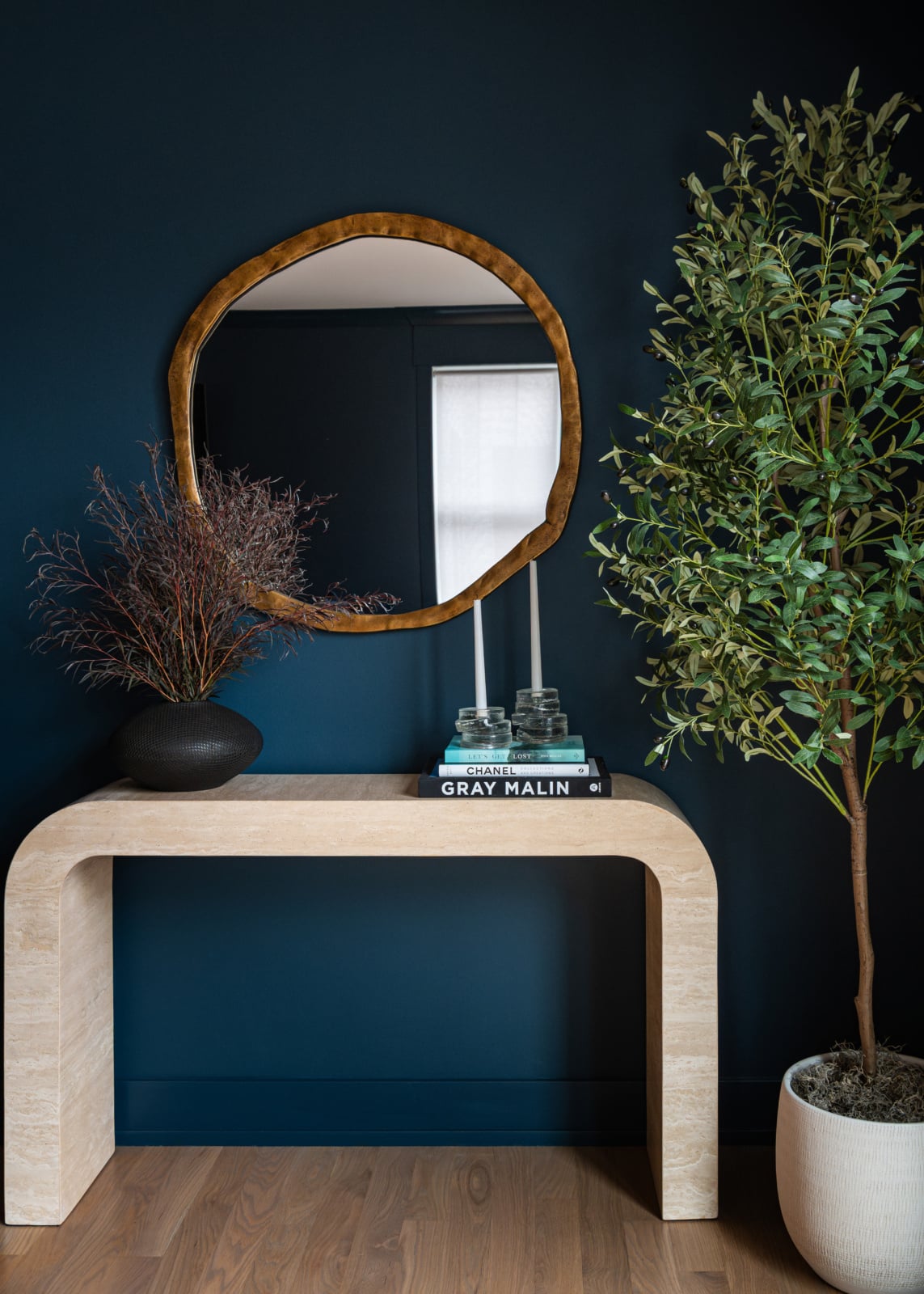A modern entryway features a curved beige console table with decor, a round wooden-framed mirror, a potted plant, and books against a dark blue wall.