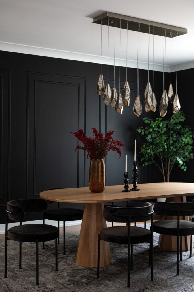 A modern dining room with a wooden oval table, black chairs, a gold vase with red flowers, two black candlesticks, a geometric chandelier, black walls, and a green plant in the corner.