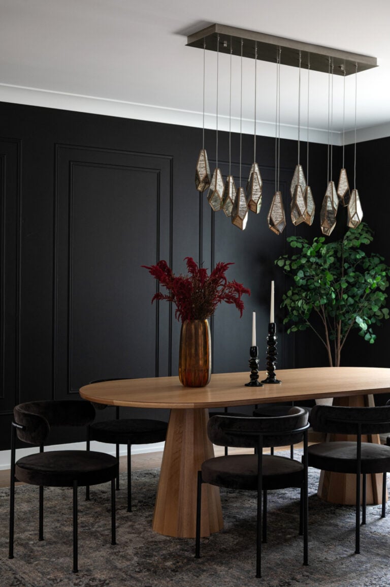A modern dining room with a wooden oval table, black chairs, a gold vase with red flowers, two black candlesticks, a geometric chandelier, black walls, and a green plant in the corner.
