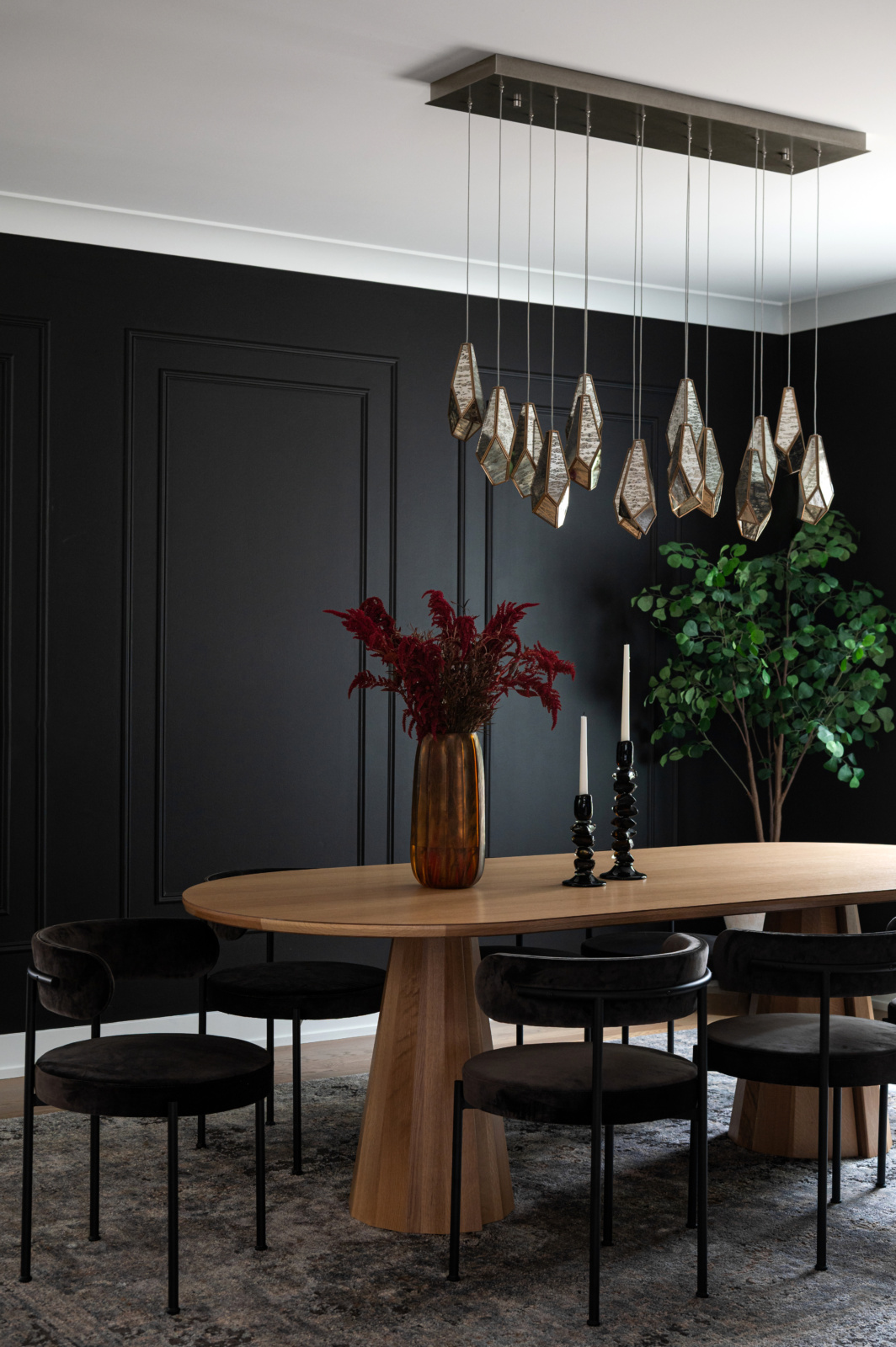 A modern dining room with a wooden oval table, black chairs, a gold vase with red flowers, two black candlesticks, a geometric chandelier, black walls, and a green plant in the corner.