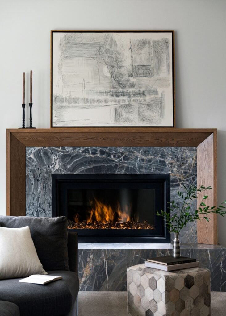 A modern living room features a marble fireplace with a wooden frame, a burning fire, abstract artwork above, and a sofa with cushions and a small side table with a vase.