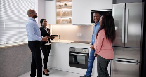 Four people stand and talk in a modern kitchen with white cabinets, a built-in oven, microwave, and a stainless steel refrigerator.