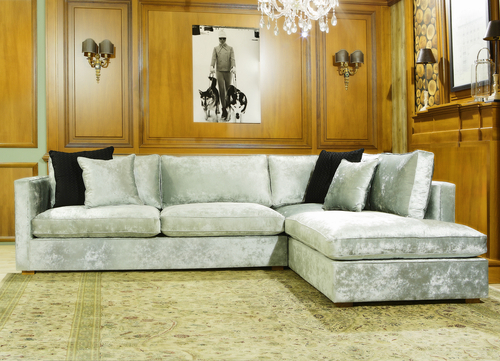 Modern silver velvet sectional sofa with black accent pillows on a patterned rug, set against wood-paneled walls with framed artwork and wall sconces.