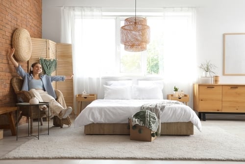A woman sits smiling on a chair in a bright, modern bedroom with a double bed, light wood furniture, and large window letting in natural light.