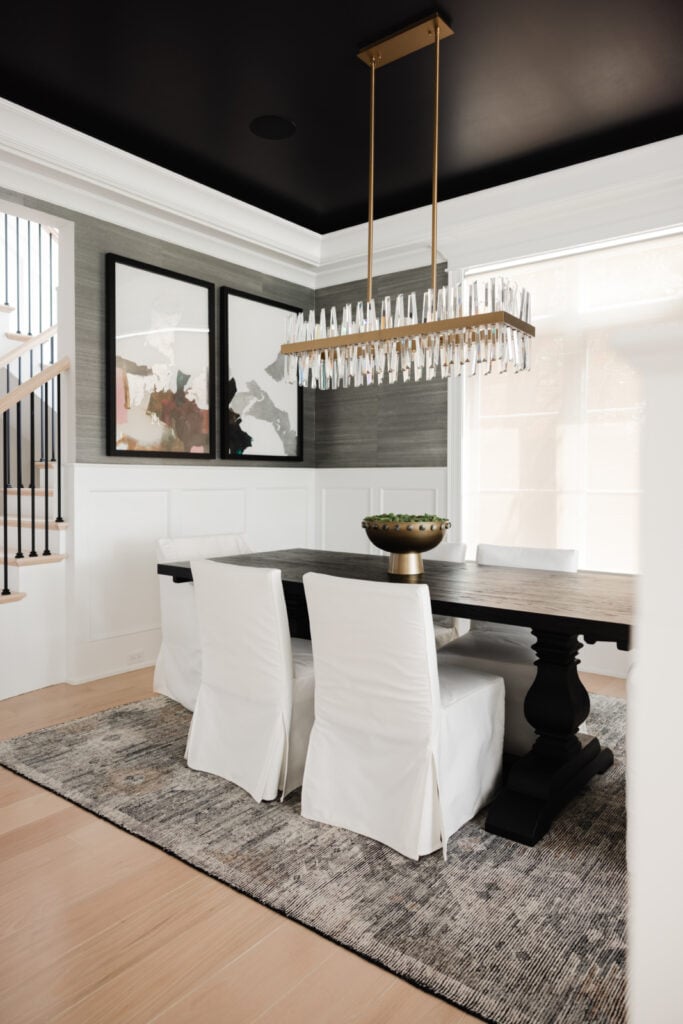 Modern dining room with a dark wood table, six white upholstered chairs, abstract wall art, a large chandelier, and a gray rug on light wood flooring.