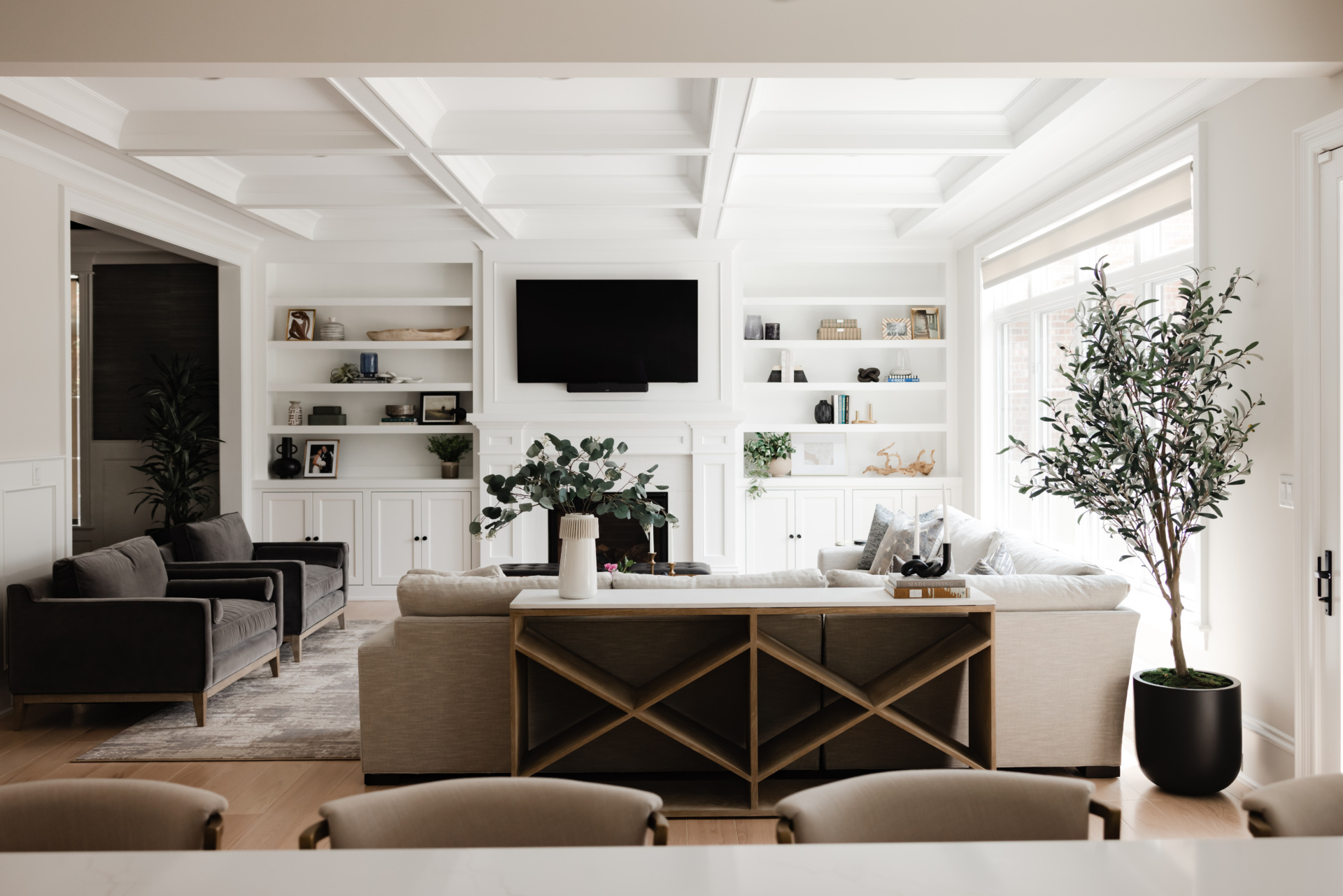 A modern living room with neutral-toned furniture, built-in shelves, a wall-mounted TV, large windows, and a potted plant near the sofa.