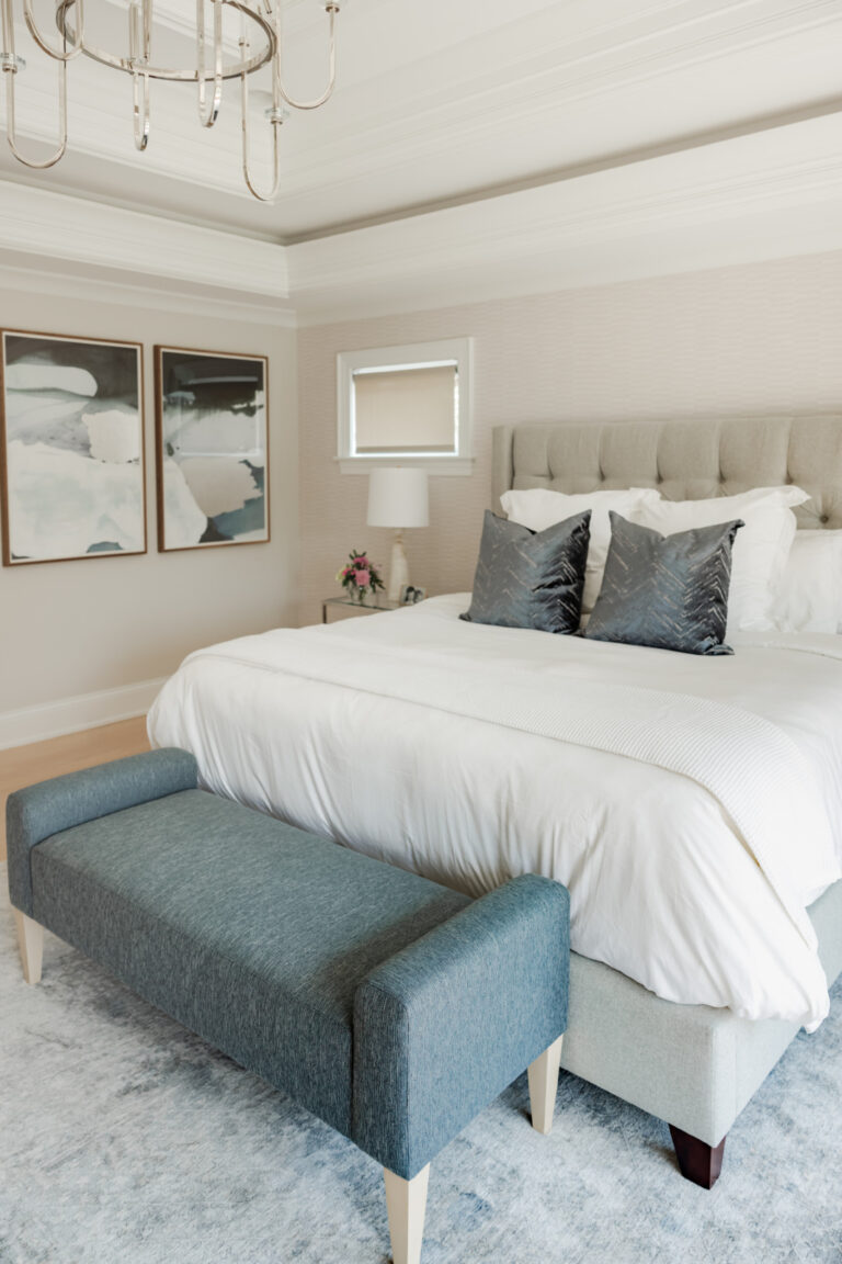 A modern bedroom with a large bed, two gray pillows, a blue upholstered bench at the foot, two abstract paintings, and a chandelier overhead.