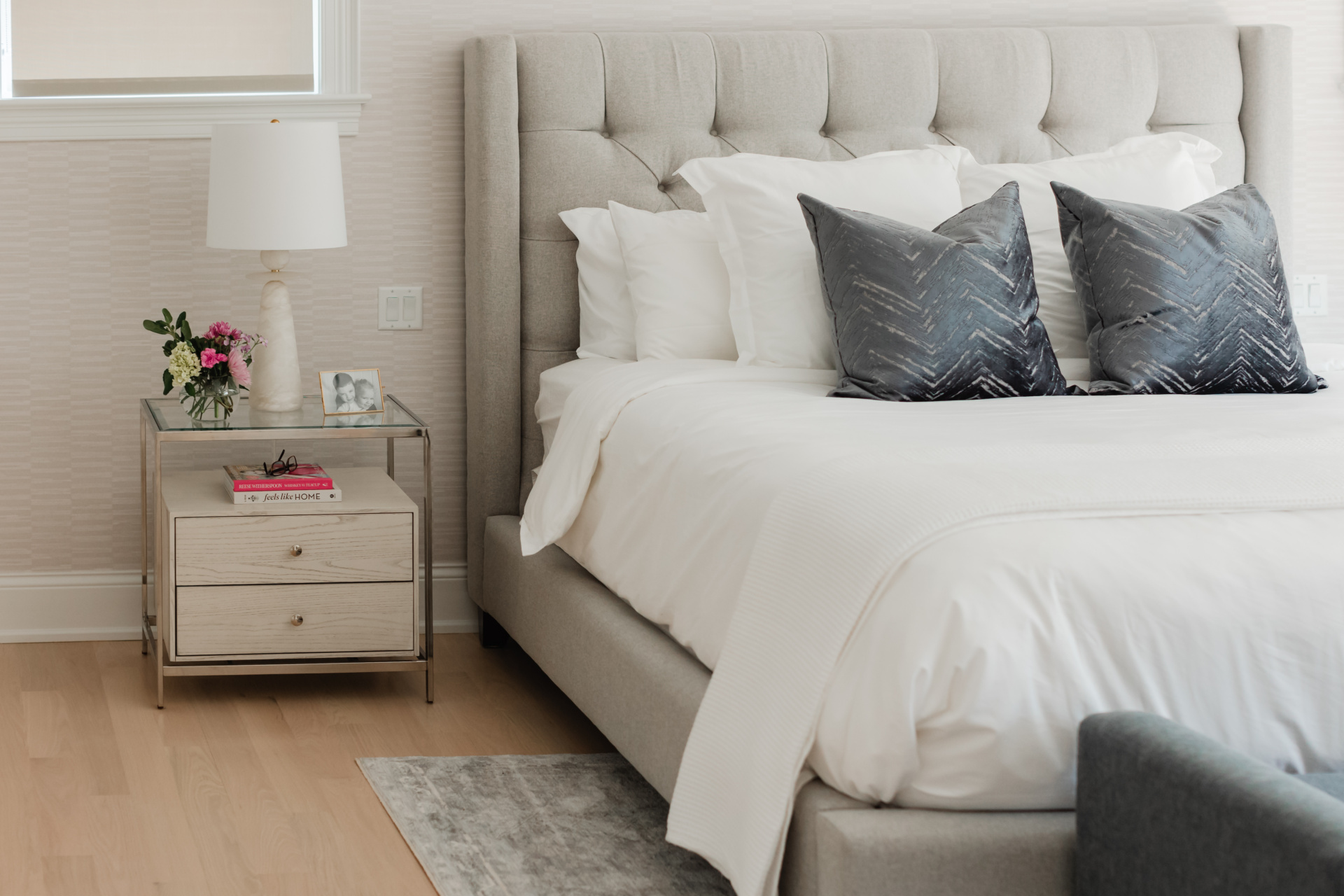 A modern bedroom with a grey upholstered bed, white bedding, two dark accent pillows, and a glass nightstand holding a lamp, flowers, a framed photo, and a book.