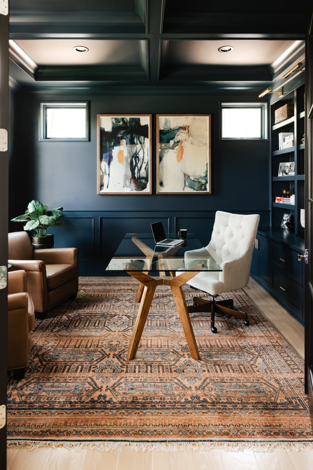 A modern home office with dark walls, a glass desk, tufted chair, leather armchair, decorative rug, potted plant, shelves, and abstract art on the wall.