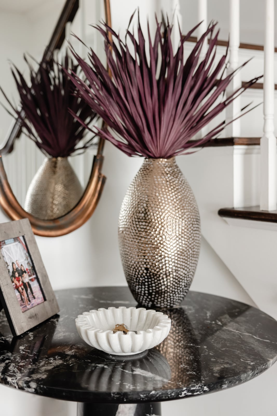 A marble-topped table holds a textured metallic vase with purple leaves, a framed photo, and a white decorative bowl; a staircase and mirror are in the background.