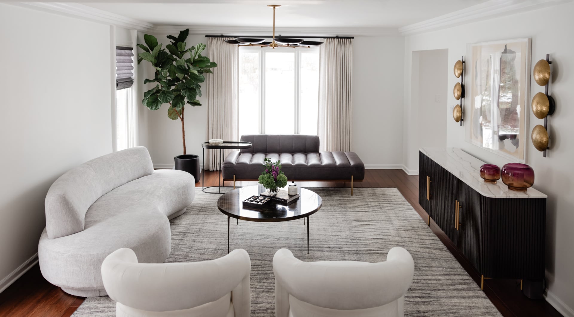 Modern living room with white curved sofa, two white chairs, a black chaise lounge, round coffee table, large rug, potted plant, and wall art with decorative sconces above a dark credenza.