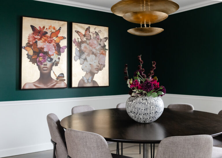 A dining room with a dark round table, light gray chairs, a large floral vase, gold pendant lights, and two framed art pieces of women with flowers and butterflies in their hair.