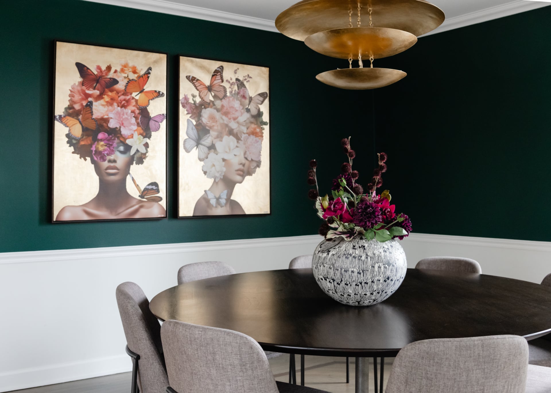A dining room with a dark round table, light gray chairs, a large floral vase, gold pendant lights, and two framed art pieces of women with flowers and butterflies in their hair.