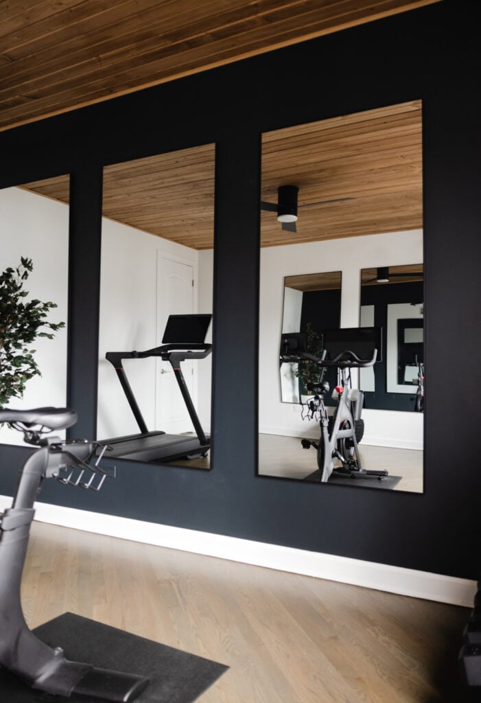 A treadmill and stationary bike are reflected in two large mirrors on a black accent wall in a modern home gym with wood flooring and ceiling.
