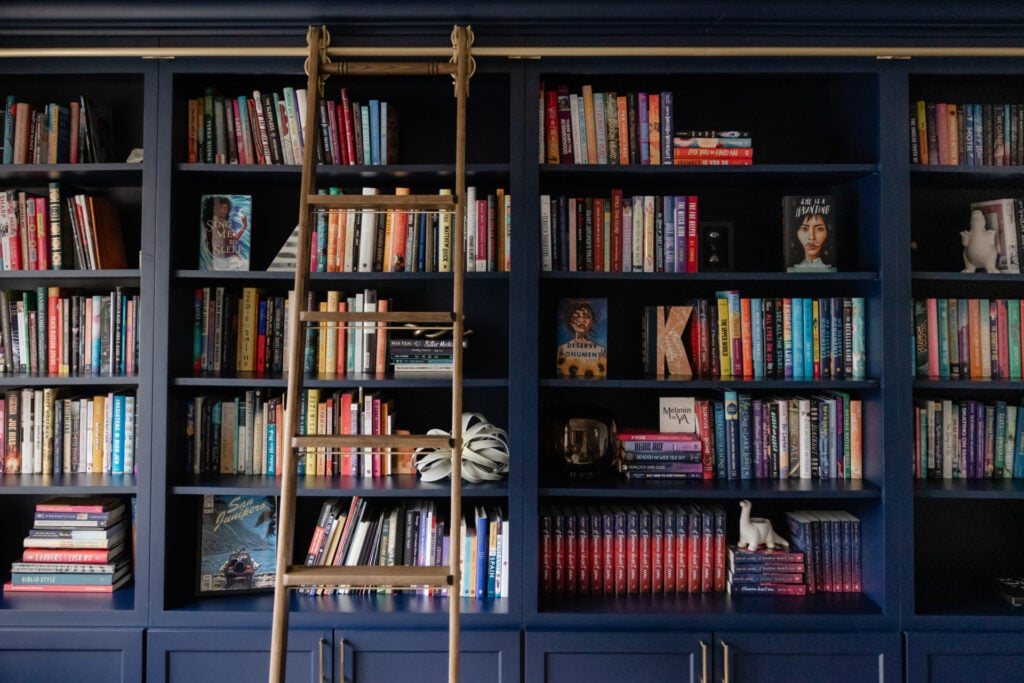 A dark blue built-in bookshelf filled with assorted books, decorative objects, and a rolling ladder positioned in front.